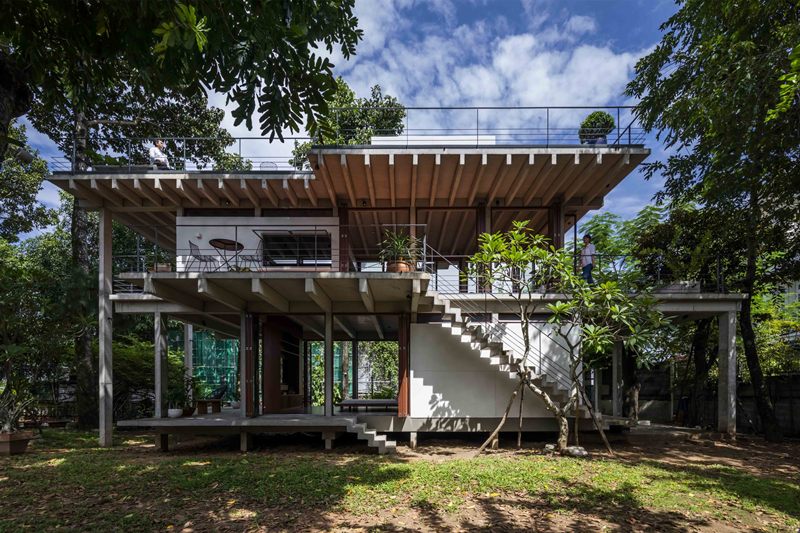 Foto rumah panggung dua lantai berbahan beton dengan fasad simple dan jendela lebar, dikelilingi pepohonan kecil.