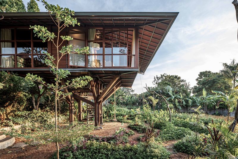 Ilustrasi rumah panggung kayu dengan balkon terbuka, dikelilingi tanaman tropis rimbun.