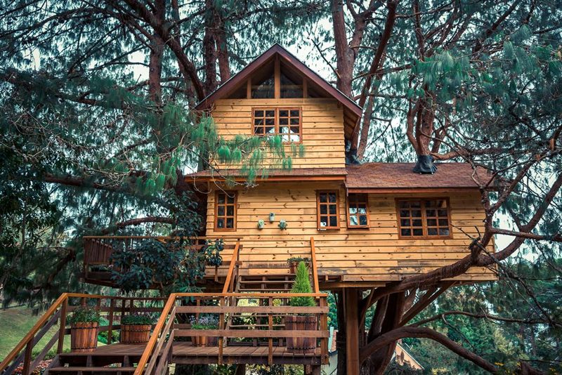 Perspektif rumah kayu bertingkat dengan struktur seperti menggantung, dilengkapi tangga luar.