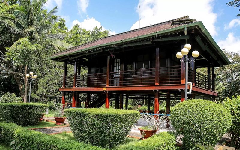 Rumah panggung dengan atap genteng tradisional, jendela kaca lebar, dan teras tropis.