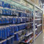 Interior toko bangunan di Kalimantan Timur dengan rak-rak penuh stok bahan bangunan seperti semen, cat kaleng, papan kayu, dan pipa PVC. Tampak juga alat-alat konstruksi seperti palu, bor listrik, gerinda, serta mesin-mesin berat seperti beton mixer dan genset yang siap pakai. Toko terlihat terorganisir dengan stok lengkap dan siap melayani kebutuhan proyek.