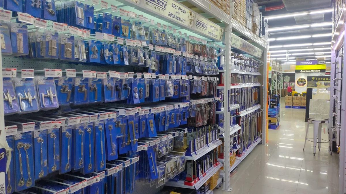 Interior toko bangunan di Kalimantan Timur dengan rak-rak penuh stok bahan bangunan seperti semen, cat kaleng, papan kayu, dan pipa PVC. Tampak juga alat-alat konstruksi seperti palu, bor listrik, gerinda, serta mesin-mesin berat seperti beton mixer dan genset yang siap pakai. Toko terlihat terorganisir dengan stok lengkap dan siap melayani kebutuhan proyek.